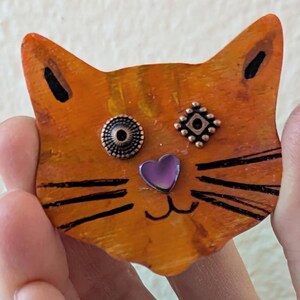 May include: Orange wooden cat figurine with black painted whiskers and a purple heart nose. The cat has two copper-colored buttons for eyes, one with a circular design and the other with a square design.