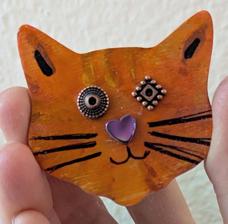 May include: Orange wooden cat figurine with black painted whiskers and a purple heart nose. The cat has two copper-colored buttons for eyes, one with a circular design and the other with a square design.