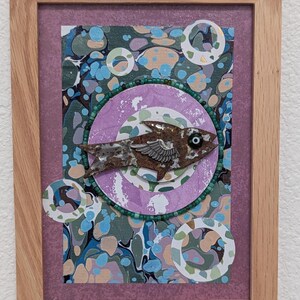 May include: A framed mixed-media artwork featuring a central brown fish cutout with a silver wing and a single eye bead. The fish is surrounded by a green beaded circle on a purple background with marbled blue, green, and tan paper accents.