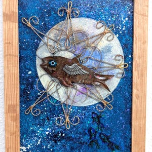 May include: A framed mixed-media artwork featuring a brown fish with small wings, set against a blue starry background with a large, pale moon. The fish is surrounded by ornate wire detailing. Additional blue fish are in the lower right corner.