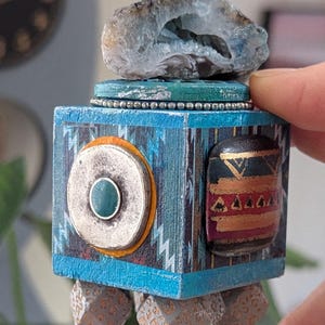May include: A handcrafted, geometric sculpture with a blue and turquoise color scheme. The sculpture features a geode, a turquoise accent, and a patterned design. The base has small, cube-shaped feet. The sculpture is held by a hand.