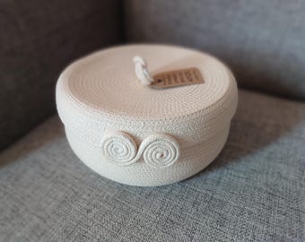 Può includere: Una scatola rotonda in corda intrecciata color crema con coperchio abbinato. Il coperchio ha una piccola etichetta attaccata con uno spago. Due spirali decorative adornano la parte anteriore della scatola. La scatola è su una superficie grigia.