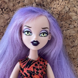 Könnte beinhalten: Eine Puppe mit lila Haaren, gelben Augen und dunkelviolettem Lippenstift. Sie trägt ein schwarz-oranges gemustertes Oberteil.