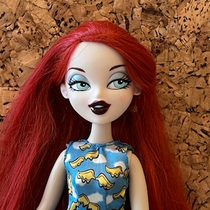 Könnte beinhalten: Puppe mit langem, leuchtend rotem Haar und heller Haut. Die Puppe hat auffällige blaue Augen, dunklen Lippenstift und trägt ein blaues Kleid mit Wolken- und Flügelmuster. Der Hintergrund ist eine strukturierte, braune Oberfläche.