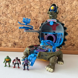 Op de afbeelding: Een groene, bruine en blauwe Teenage Mutant Ninja Turtles speelset met een speelgebied in de vorm van een schildpadden schild met een ladder, een klein kanon en een geheim vak. Vier kleine action figuren zijn inbegrepen: Leonardo, Donatello, Raphael en Michelangelo.