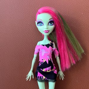 Peut inclure: Une poupée à la peau verte avec des cheveux roses et verts portant une robe noire et rose à motifs dentelés.