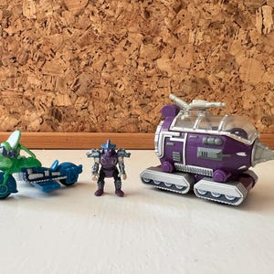 Op de afbeelding: Een verzameling speelgoedvoertuigen en figuren. Inclusief een groene en blauwe motorfiets, een paarse en zilveren figuur en een paarse boortank met een doorzichtig dak. Het speelgoed staat tegen een achtergrond van kurk.