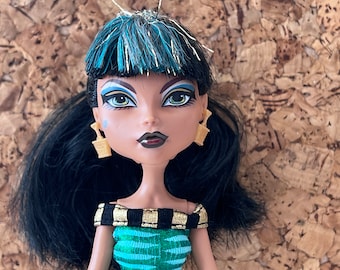 May include: A black-haired doll with blue and silver hair accents. She is wearing a green and gold dress and gold earrings. Her eyes are green and her lips are dark red.