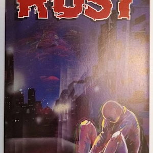 May include: A comic book cover with the title "RUST" in red block letters. The cover art depicts a person sitting on a rooftop in a city at night. The person is wearing a blue shirt and pants. The city skyline is visible in the background.