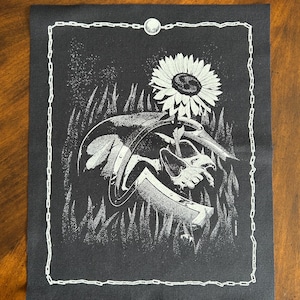 May include: Black and white graphic art print featuring a sunflower, a shield, and a hand holding a sword. The artwork is framed with a white chain-link border and a moon at the top. The image is on a dark background.