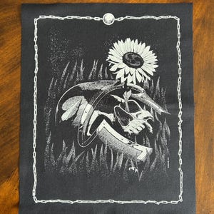 May include: Black and white graphic art print featuring a sunflower, a shield, and a hand holding a sword. The artwork is framed with a white chain-link border and a moon at the top. The image is on a dark background.