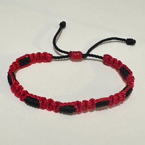 May include: Red and black macrame bracelet with an adjustable sliding knot closure. The bracelet features a repeating pattern of red and black knots. The adjustable closure is made of black cord.