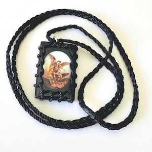 May include: A black pendant necklace with a religious image of an angel. The rectangular pendant is framed in black and suspended from a thick, black rope-style cord. The image shows a winged figure in gold, brown, and white tones.