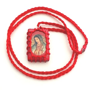 May include: A red pendant necklace featuring a rectangular pendant with an image of a religious figure. The pendant is framed in red and attached to a red woven cord. The image depicts a woman with a blue veil and gold accents.