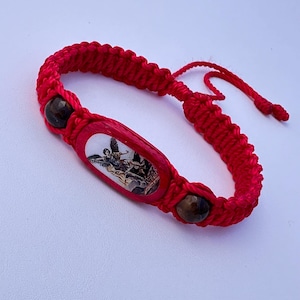 Puede incluir: Pulsera trenzada roja con un dije ovalado de madera con una imagen de un ángel. La pulsera tiene cuentas marrones de ojo de tigre.