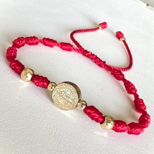 May include: Red woven bracelet with gold-colored beads and a gold medallion. The medallion features an embossed design. The bracelet is adjustable with a sliding knot closure. The bracelet is displayed on a white surface.