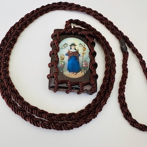 Puede incluir: Un collar con colgante religioso que presenta un colgante rectangular con una imagen de un santo con túnica azul. El colgante está enmarcado con cuerda marrón oscuro y suspendido de un collar de cordón trenzado a juego.