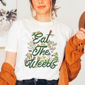 May include: White t-shirt with a green graphic of flowers and the text "Eat the Weeds".