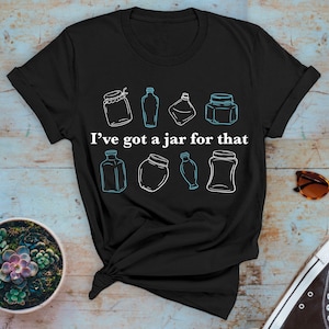 Puede incluir: Camiseta negra con un contorno blanco de varios frascos y botellas. El texto "Tengo un frasco para eso" está impreso en blanco en la camiseta.