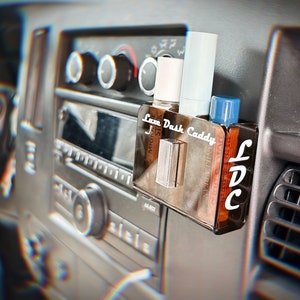 May include: A clear plastic car dash caddy with the text "Luxe Dash Caddy" printed on the front. The caddy is holding two small bottles and a tube of lip balm. The caddy is mounted on the dashboard of a car.