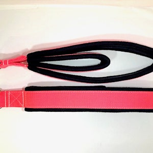 May include: A pink and black exercise strap with a metal ring on each end. The strap is made of a soft, padded material and is designed for stretching and resistance training.