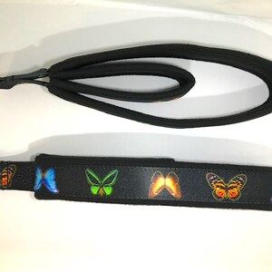May include: Black dog leash and matching wrist strap. The leash features a padded handle and a metal ring for easy attachment. The wrist strap is black with colorful butterfly graphics. The butterflies are orange, blue, and green.