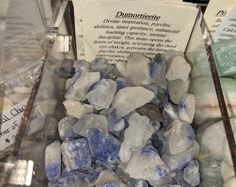 May include: A clear plastic container filled with raw dumortierite crystals. The crystals are a mix of white and deep blue, with a rough, natural texture. A small card with text about the crystals is visible. The crystals are labeled "Dumortierite in quartz".