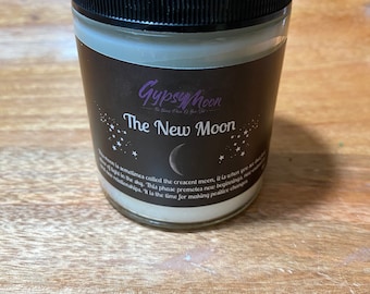 May include: A candle in a clear glass jar with a black lid. The label is dark brown with the text "Gypsy Moon" and "The New Moon" in purple and white. The candle is on a wooden surface.