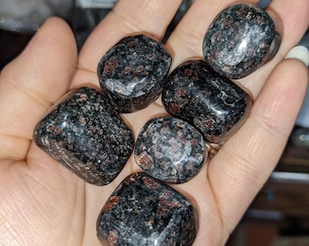 May include: A collection of polished, dark-colored stones with speckled red and brown inclusions. The stones are held in a hand, showcasing their smooth, rounded shapes and natural variations. The stones appear to be gemstones or crystals.