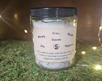 May include: A clear glass jar with a black lid, containing a candle with a label that reads "Money, Success, Wealth, Joy, Happiness." The candle is on a bed of green moss. The label also states the candle is made with oils, herbs, and crystals.