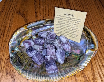 May include: A collection of purple Lepidolite crystals in a shell. The crystals are tumbled and have a smooth, polished finish. The text on the card reads: "Lepidolite If effective in calming frayed nerves, helping one to release stress and worry, and sets you on the path of willing acceptance. Provides energy of enlightened awareness so one can handle the situation in the highest way. A stone of serenity. It encourages one to respond to hostility without putting up defenses, and find the path of harmonious action and to see problems as opportunities to learn. A stone of spiritual purification and meditation."