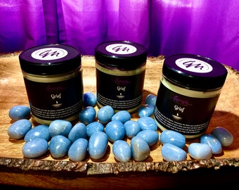 May include: Three glass jars of candle with black labels that read "Grief" and "Gypsy Moon" with a candle graphic. The jars are on a wooden surface with blue stones scattered around them. The background is a purple curtain.