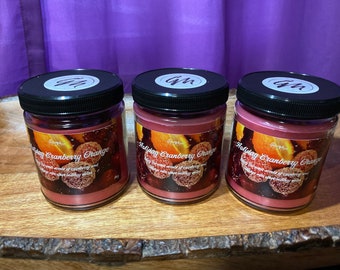 May include: Three glass jars with black lids containing red scented candles. The candles are labeled "Holiday Cranberry Orange" and have a festive orange and cranberry design.