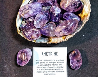 May include: A collection of polished ametrine stones, a natural combination of amethyst and citrine, displayed in a decorative shell. The stones are a deep purple color. A white card with the word "AMETRINE" and description is also present.