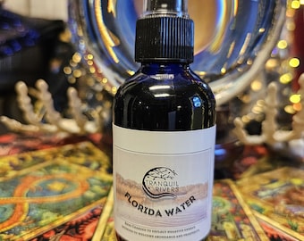 May include: A dark blue glass bottle of Florida Water with a black spray top. The label reads "Tranquil Rivers Florida Water" with text below. The bottle is in front of a large glass sphere and a colorful patterned surface.