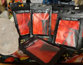 May include: Several black pouches with the text "Coffin Nails" are displayed. Each pouch contains black nail tips and a red ribbon against a red background. A clear crystal skull is visible in the background.