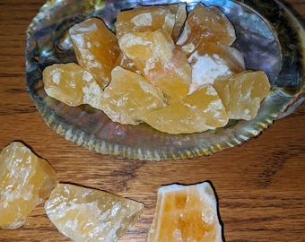 May include: A collection of rough, golden calcite crystals displayed in an iridescent abalone shell. Several additional crystals are scattered around the shell on a wooden surface. The crystals have a translucent, honey-like appearance.