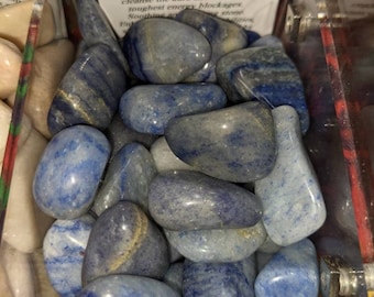 May include: A collection of polished blue quartz stones in a clear container. The stones vary in shades of blue and gray, with some displaying streaks of lighter color. A white label reads "Blue Quartz" and describes its properties.