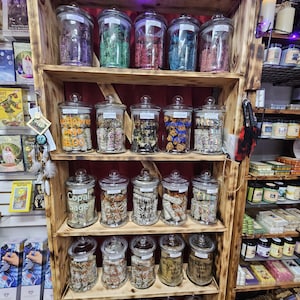 May include: A wooden shelf unit filled with glass jars containing various types of sage, white sage, copal sage, and other herbs. The jars are labeled with the type of sage and the price. The shelf is decorated with fairy lights and is located in a shop.