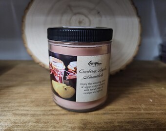 May include: A glass jar candle with a black lid, labeled "Cranberry Apple Marmalade." The label features an illustration of apples and jars of jam. The candle has a pink hue and sits on a wooden surface.