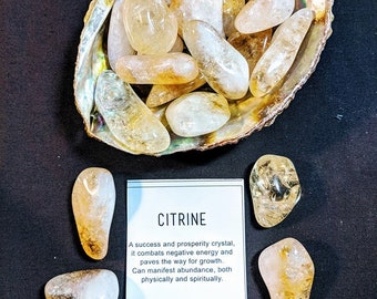 May include: A collection of polished citrine stones in shades of yellow and gold. The stones are displayed in a natural shell and arranged around a card that reads "CITRINE". The card describes citrine as a success and prosperity crystal.