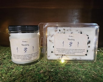 May include: Two candles, one in a glass jar with a black lid and the other in a clear plastic container. Both have labels that read "Healing" with a symbol and text. The candles are on a bed of green moss.