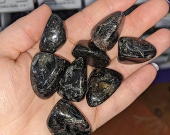 May include: A handful of polished black stones with reflective, metallic inclusions. The stones are smooth and rounded, varying in size and shape. The background shows small containers with other stones.