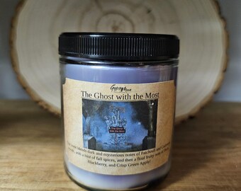 May include: A glass jar candle with a black lid, labeled "The Ghost with the Most." The label features an illustration and scent notes. The candle has a light purple wax and is on a wooden surface.