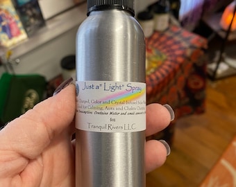 May include: A silver spray bottle with a black cap is held in a hand. The label reads "Just a Light Spray" and lists ingredients and the brand "Tranquil Rivers LLC." The bottle contains 4 oz of liquid.