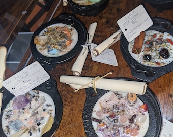 May include: Assortment of small cauldron-shaped candles with various crystals, herbs, and other ingredients. The candles are labeled with names and prices, such as "Anti-Anxiety Candle Cauldron $22", "Letting Go Charm Candle Cauldron $22", and "Justice Candle Cauldron $22".