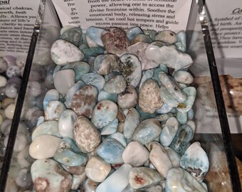 May include: A display of Larimar stones in a clear container. The stones are a mix of light blue, white, and brown, with a smooth, polished appearance. A sign provides information about the stones.