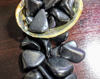 May include: A collection of polished, black hematite stones displayed in an iridescent, natural seashell. The stones are smooth and reflective, with a dark, metallic sheen. The seashell has a textured, multi-colored surface, and the background is a dark wood.