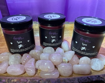 May include: Three jars of Gypsy Moon Love candles with black labels and black lids, surrounded by rose quartz stones. The labels feature a heart, an illustration of Cupid, and the text "Love". The candles are on a wooden tray.
