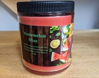 May include: A glass jar candle with a black lid and a red label that says "Watermelon Mint". The label has a picture of watermelon slices, lemons, and mint leaves. The text on the label reads: "Refresh your senses with the amazing mouthwatering scent of watermelon with a hint of mint!"